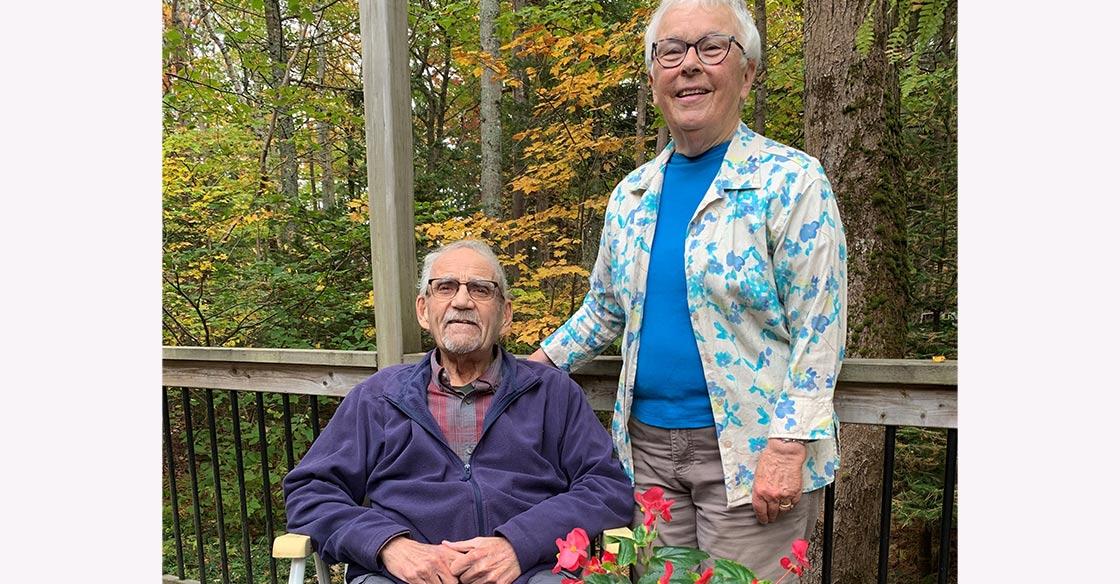 An elderly couple stands on a porch, surrounded by fall colours. The husband, Bob,  sits in a chair on the left to his wife, Wendy, who's standing next to him.