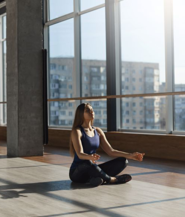 A woman sits in a yoga pose, facing large glass windows. 