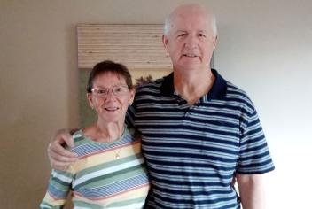 Older couple stands together in front of beige wall and painting