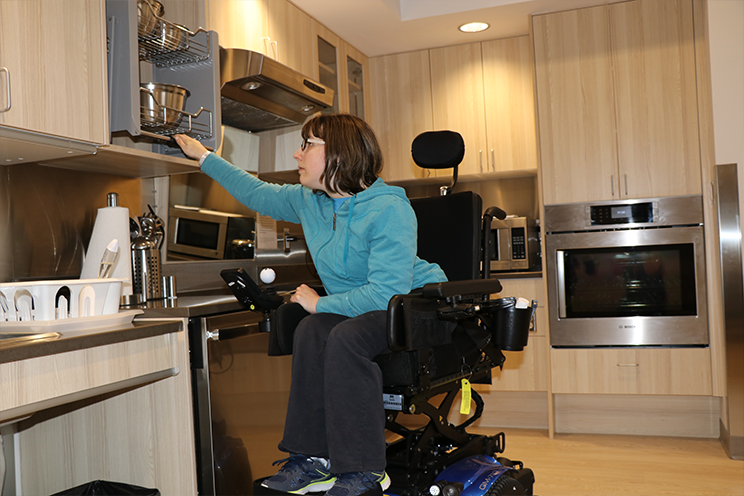 A rehab patient in a wheel chair is adjusting the cupboard height to reach the dishes at the Independent Living Simulation Suites