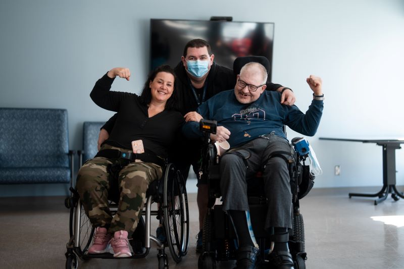 Two rehabilitation patients, seated in wheelchairs, and a therapist, smile at the camera. 