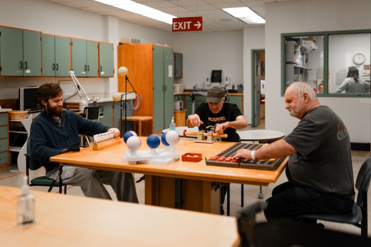 Three men sitting at a table are using equipment and games in a physical rehabilitation space.