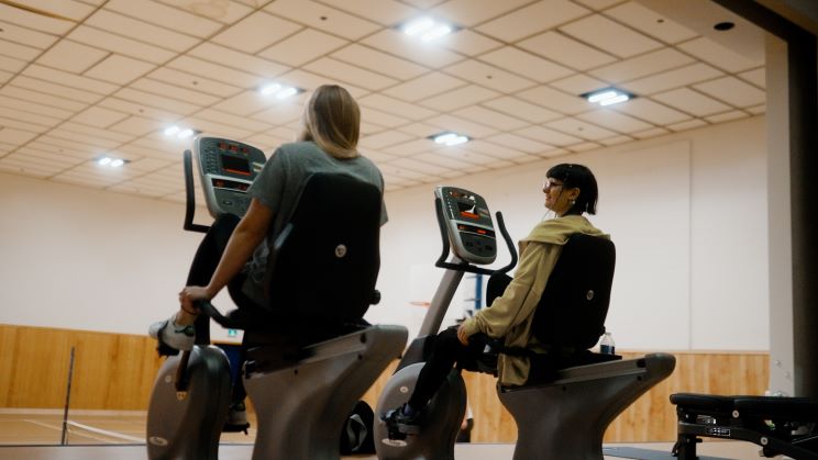 Two people are using exercise bikes side by side in a gymnasium; they appear to be mid-conversation.