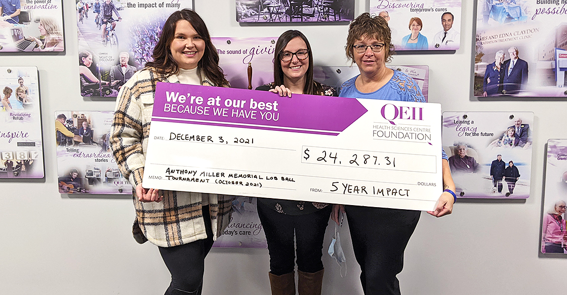Emily White, Angie White, and Caitlin Parks hold a large QEII Foundation cheque in front of a mural wall 
