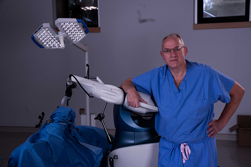 A male surgeon stands facing the camera with his arm propped on an orthopaedic surgical robot. 