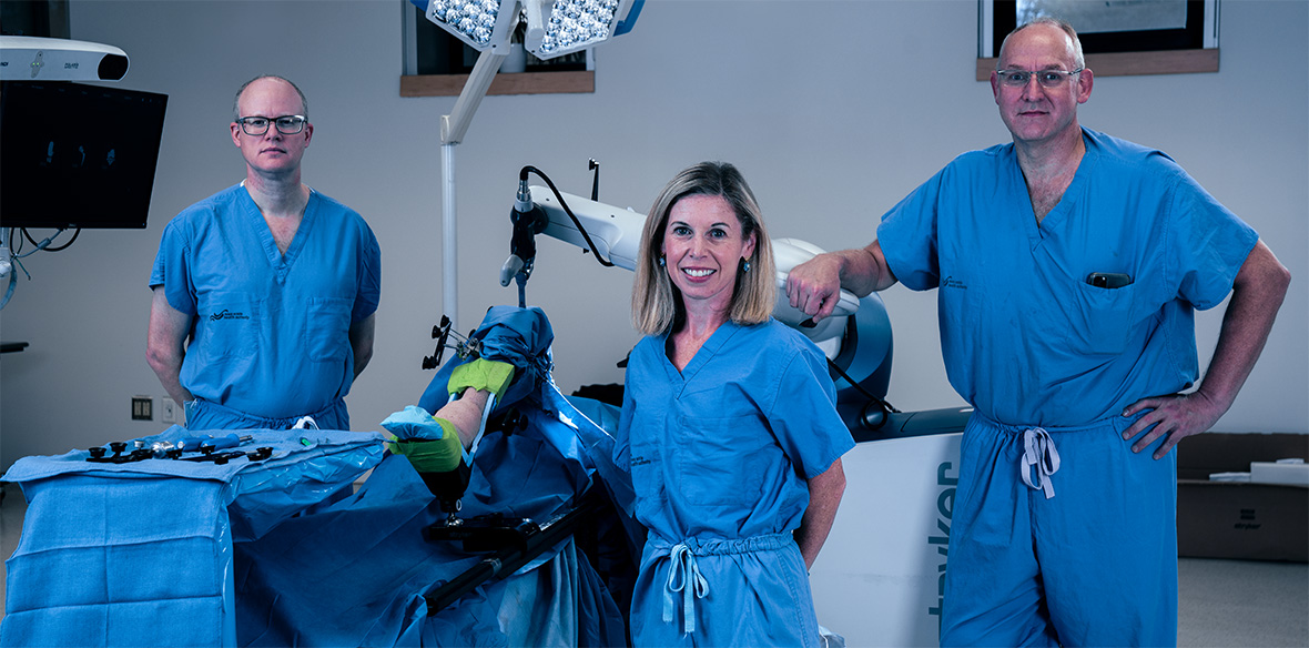 Three researchers dressed in blue scrubs stand with the orthopaedic surgical robot