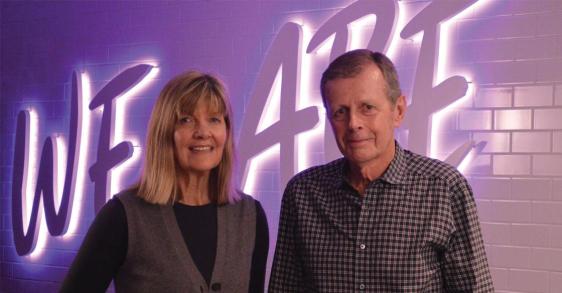 A man and woman smile at the camera, standing in a white wall with a glowing purple sign behind them that reads We Are