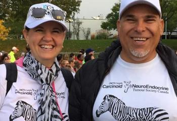 Jackie stands alongside her husband, Joe, at a busy, outdoor event. Both are smiling and wearing white t-shirts with zebra graphics