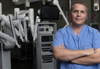 A patient wearing blue hospital scrubs stands in an OR with a surgical robot behind him. His arms are crossed as he looks into the camera.