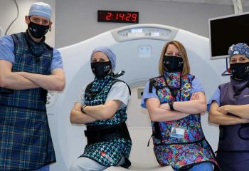 Team of four, wearing black We Are masks and lead vests, stand in front of a white CT fluoroscopy machine.