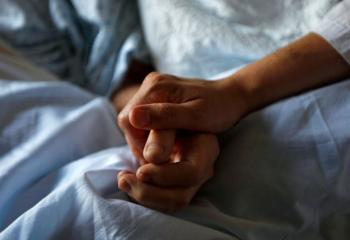 Close-up shot of two hands, holding one another for comfort and support. The hands are positioned on a blue hospital sheet, and both individuals are wearing johnny shirts 