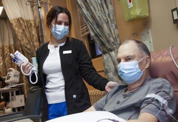 A QEII nurse stands next to an older male patient with her hand on his shoulder laying in bed receiving chemotherapy therapy treatment