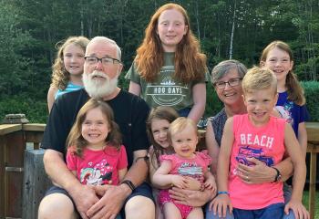 An older couple sitting on a deck surrounded by their seven smiling grandchildren