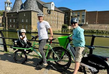 A family photograph of Dr. John Burka, his son and infant granddaughter on bright green bicycles, pictured in front of a canal and historic brick building. They are all smiling on a bright, sunny day.