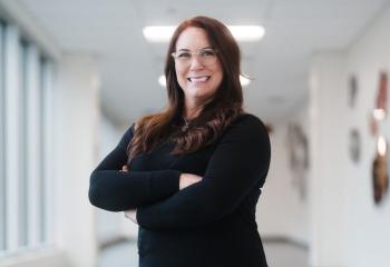 A woman with long brown hair, glasses, and dressed in black clothing stands in a brightly lit hallway, smiling at the camera with her arms crossed. 