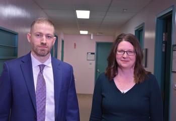 Two people, one wearing a suit, stand in a hallway, smiling. 