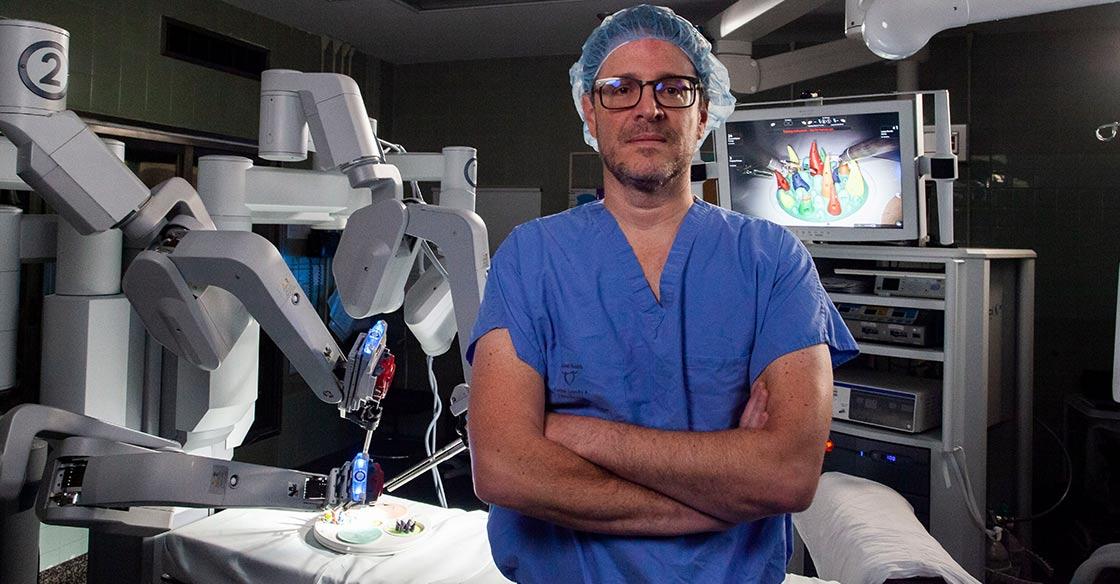 Surgeon, Dr. Ricardo Rendon, wears blue scrubs and glasses while standing in an operating room alongside the da Vinci surgical robot.
