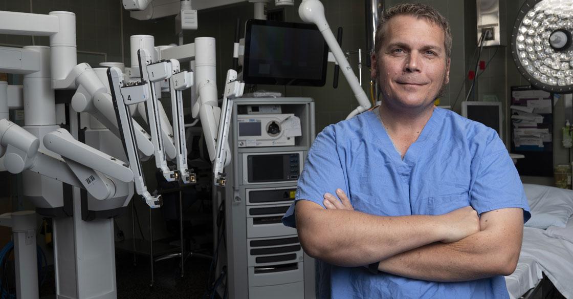 A patient wearing blue hospital scrubs stands in an OR with a surgical robot behind him. His arms are crossed as he looks into the camera.