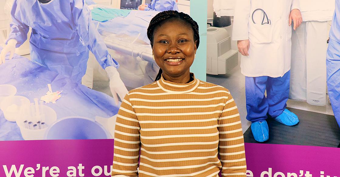 Yemisi smiles and stands proudly in front of a QEII Foundation backdrop