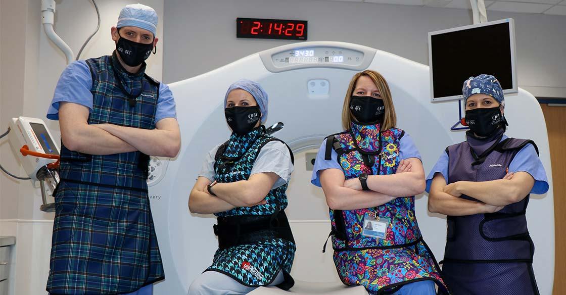 Team of four, wearing black We Are masks and lead vests, stand in front of a white CT fluoroscopy machine.