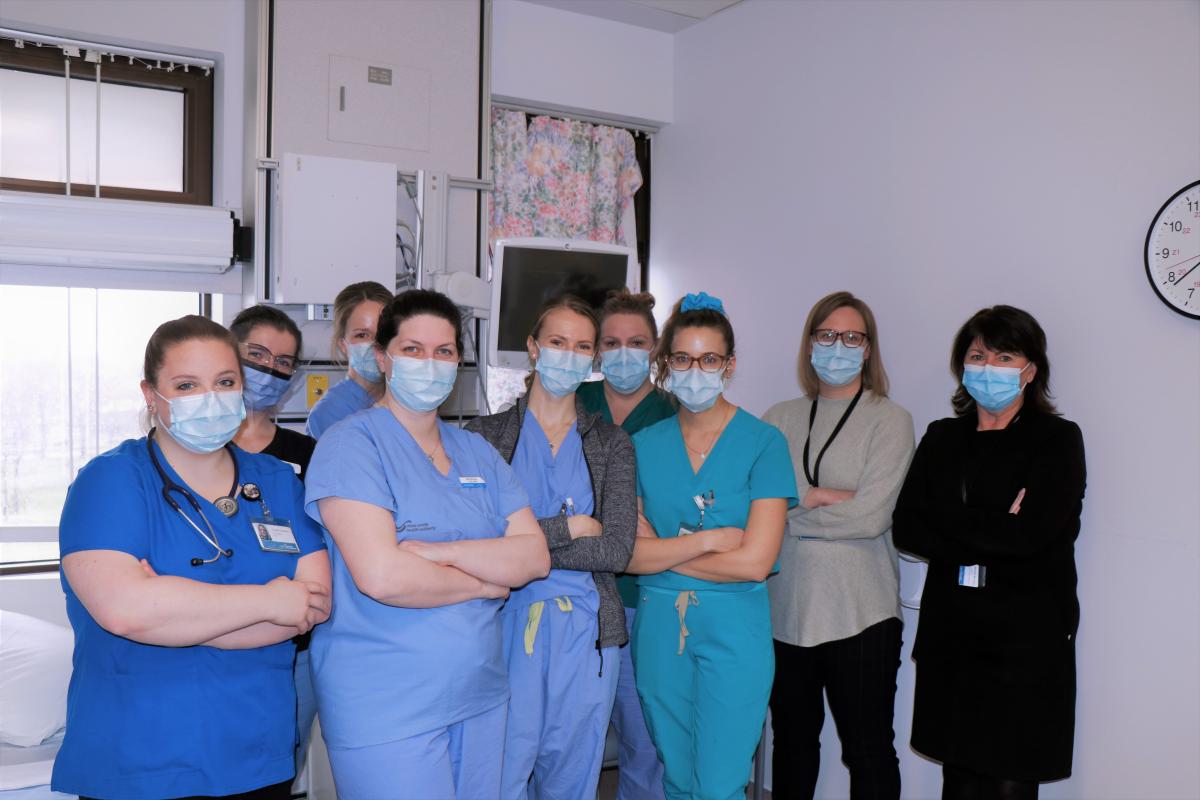CCU team, all wearing scrubs and masks, stand in a hospital room with their arms crossed