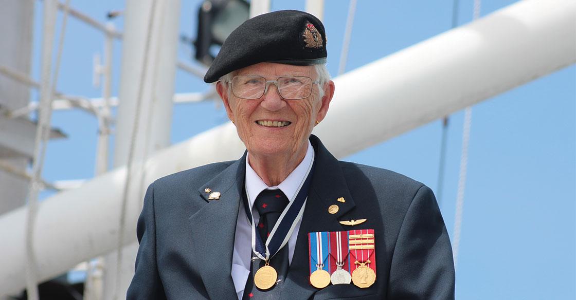 Older woman wearing her  Lieutenant-Commander uniform, stands in front of a white mast belonging to the HMCS Sackville ship