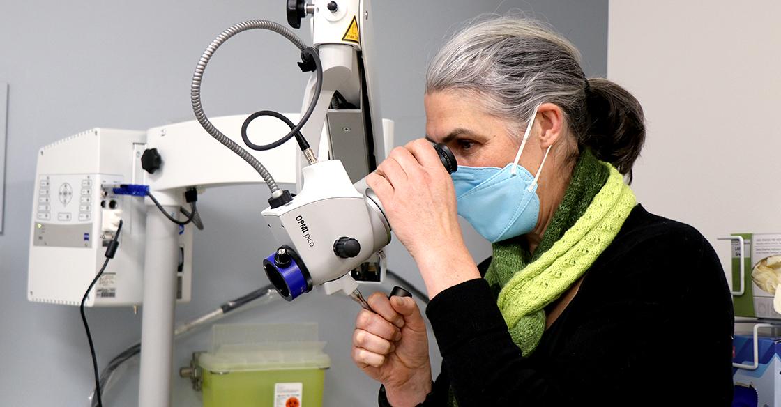 Dr. Katharina Kieser wears a green scarf and blue mask. Her side profile looks through a piece of machinery with eye lenses, known as a colposcope.