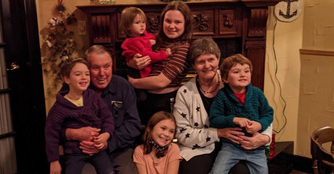 An elderly couple sitting in their living room, surrounded by five of their young grandchildren. Some of the grandchildren are in their laps, all wearing big, bright smiles
