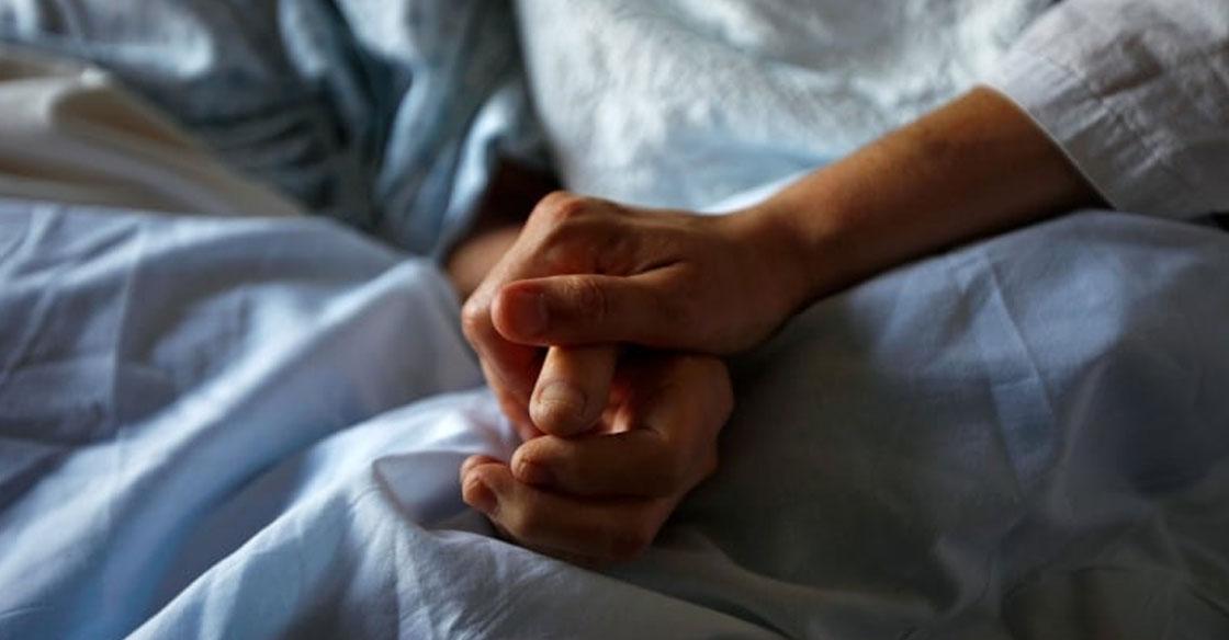 Close-up shot of two hands, holding one another for comfort and support. The hands are positioned on a blue hospital sheet, and both individuals are wearing johnny shirts