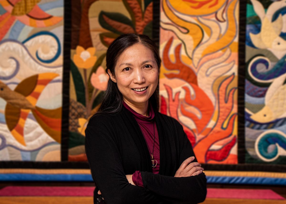 A woman, standing with her arms crossed, smiles at the camera. She stands in front of a colourful tapestry.