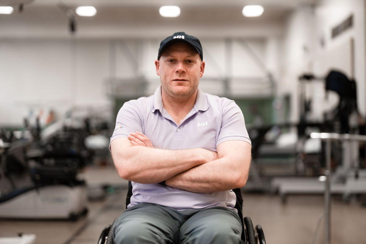 A man sitting in a wheelchair, arms crossed and smiling towards the camera. He is sitting in a gym-type setting. 