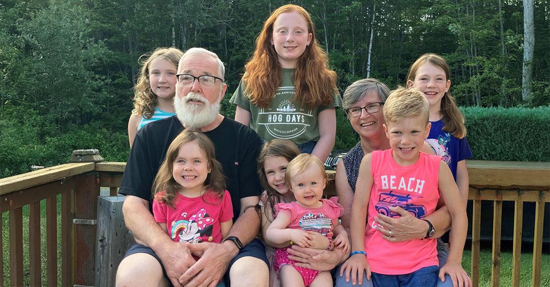 An older couple sitting on a deck surrounded by their seven smiling grandchildren