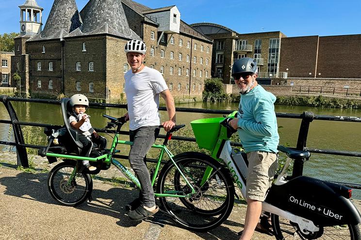 A family photograph of Dr. John Burka, his son and infant granddaughter on bright green bicycles, pictured in front of a canal and historic brick building. They are all smiling on a bright, sunny day.