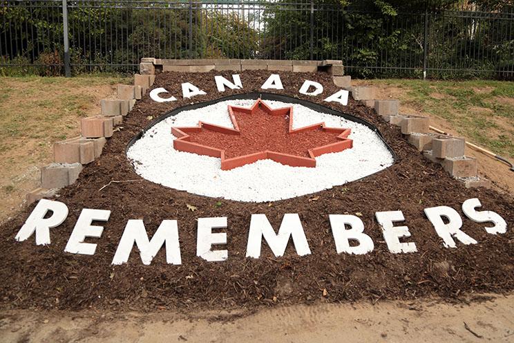 A garden bed with freshly-turned dark soil is decorated with red and white stones and mulch in the shape of a maple leaf on a white circle. Large, white, cut-out letters read 'Canada remembers' above and below the maple leaf.