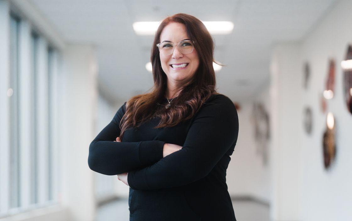 A woman with long brown hair, glasses, and dressed in black clothing stands in a brightly lit hallway, smiling at the camera with her arms crossed. 