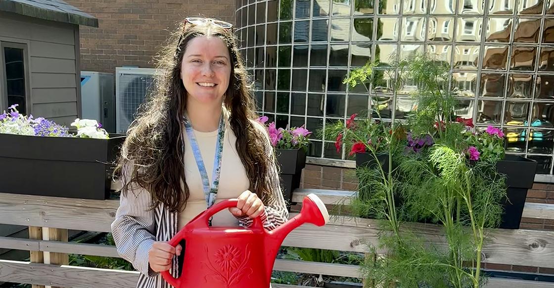 A woman with long brown hair stands smiling outdoors in a garden. In the background is a building with lots of windows. She is surrounded by flowers and plants and holds a watering can.