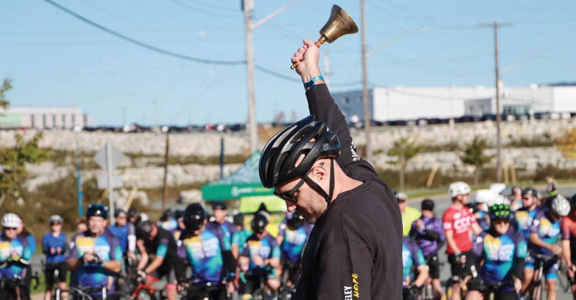 A rider wearing a black shirt and helmet rings a bell in front of a large crowd