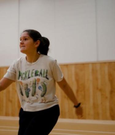 A woman holds a pickle ball racket in a gymnasium, with another woman standing in the background. 