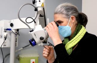 Dr. Katharina Kieser wears a green scarf and blue mask. Her side profile looks through a piece of machinery with eye lenses, known as a colposcope.