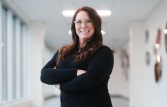 A woman with long brown hair, glasses, and dressed in black clothing stands in a brightly lit hallway, smiling at the camera with her arms crossed. 