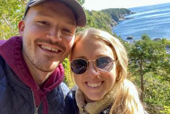 Smiling couple pose for a selfie on a hike; both are standing in front of a mountain with the ocean behind them.