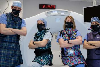 Team of four, wearing black We Are masks and lead vests, stand in front of a white CT fluoroscopy machine.