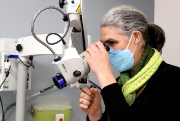 Dr. Katharina Kieser wears a green scarf and blue mask. Her side profile looks through a piece of machinery with eye lenses, known as a colposcope.