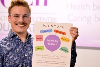 Garry Dart wears a blue patterned shirt and smiles holding up a pronouns poster