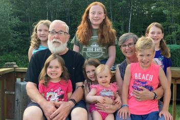 An older couple sitting on a deck surrounded by their seven smiling grandchildren